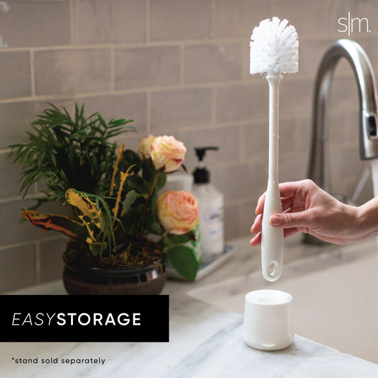 White bottle brush with fluffy bristles held in hand above matching white cylindrical storage stand on bathroom counter with marble surface, potted flowers, and subway tile backsplash.