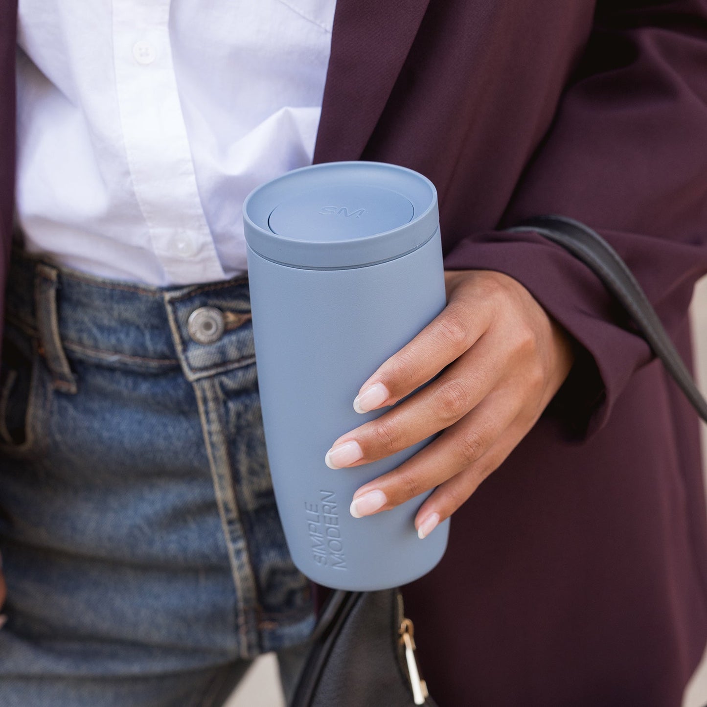 Pale blue tumbler held by person wearing deep burgundy jacket against soft blue and periwinkle background