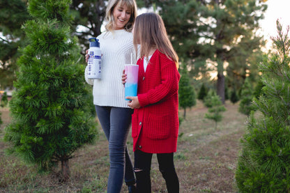 Two people standing outdoors surrounded by green evergreen shrubs, one wearing a cream-white ribbed sweater and the other in warm-toned clothing.
