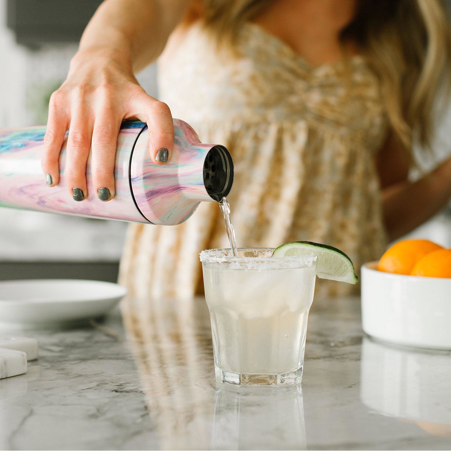 Classic stainless steel cocktail shaker with iridescent finish in blush pink and mint green, pouring liquid against soft white and gray background.