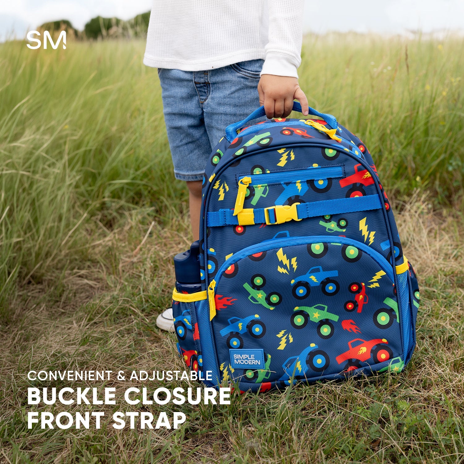 Fletcher Kids' Backpack in medium size with blue base and sports ball graphic print, displayed next to young boy in red athletic outfit holding basketball on concrete waterfront surface with cloudy sky background