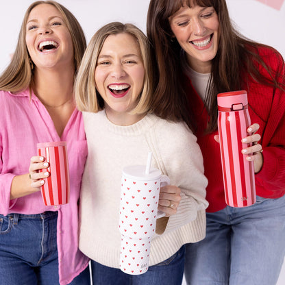 Two women in coordinating pink and red outfits holding matching red and white drinkware against a warm-toned Valentine's Day themed background.