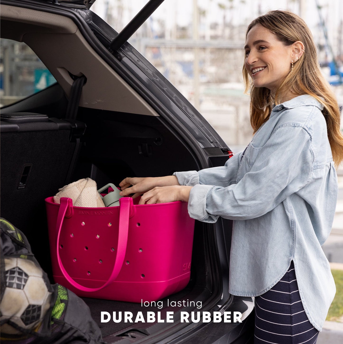 Woman in denim shirt packing a hot pink durable rubber tote bag into a car trunk in an urban setting. Text reads "long lasting DURABLE RUBBER."