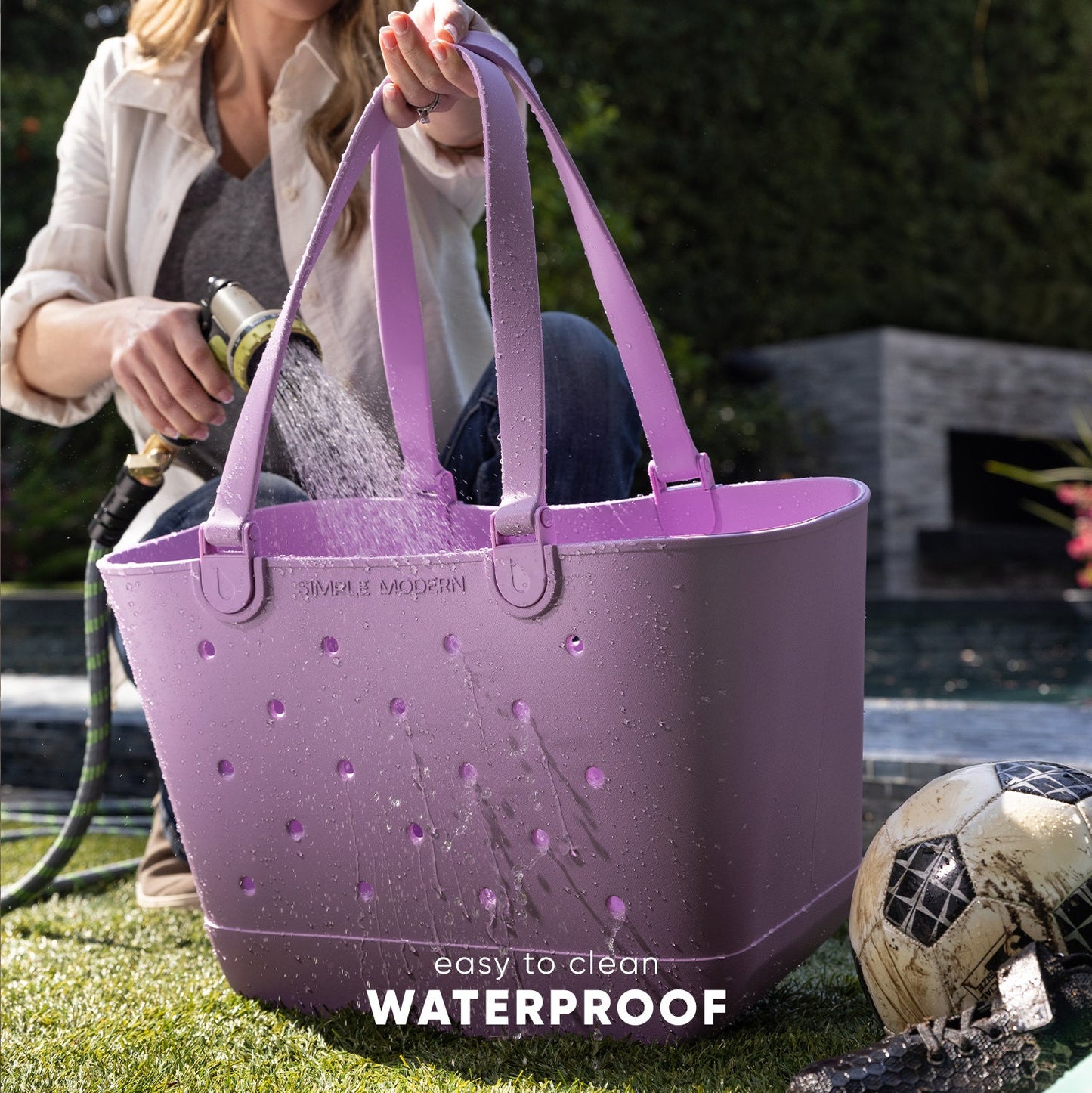Woman holding a mauve purple waterproof tote bag while rinsing it with a garden hose in a backyard, demonstrating its easy-to-clean feature. Muddy soccer ball visible on grass.