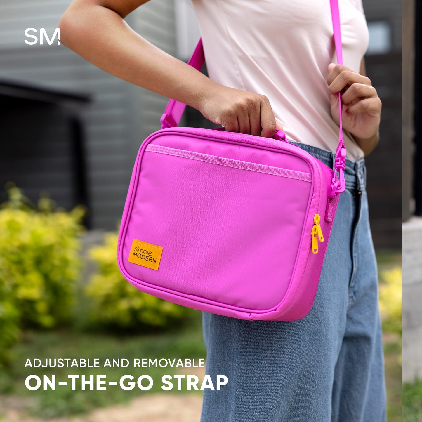 Hot pink hardshell lunch bag with adjustable shoulder strap, shown worn by person in light pink shirt and blue denim jeans