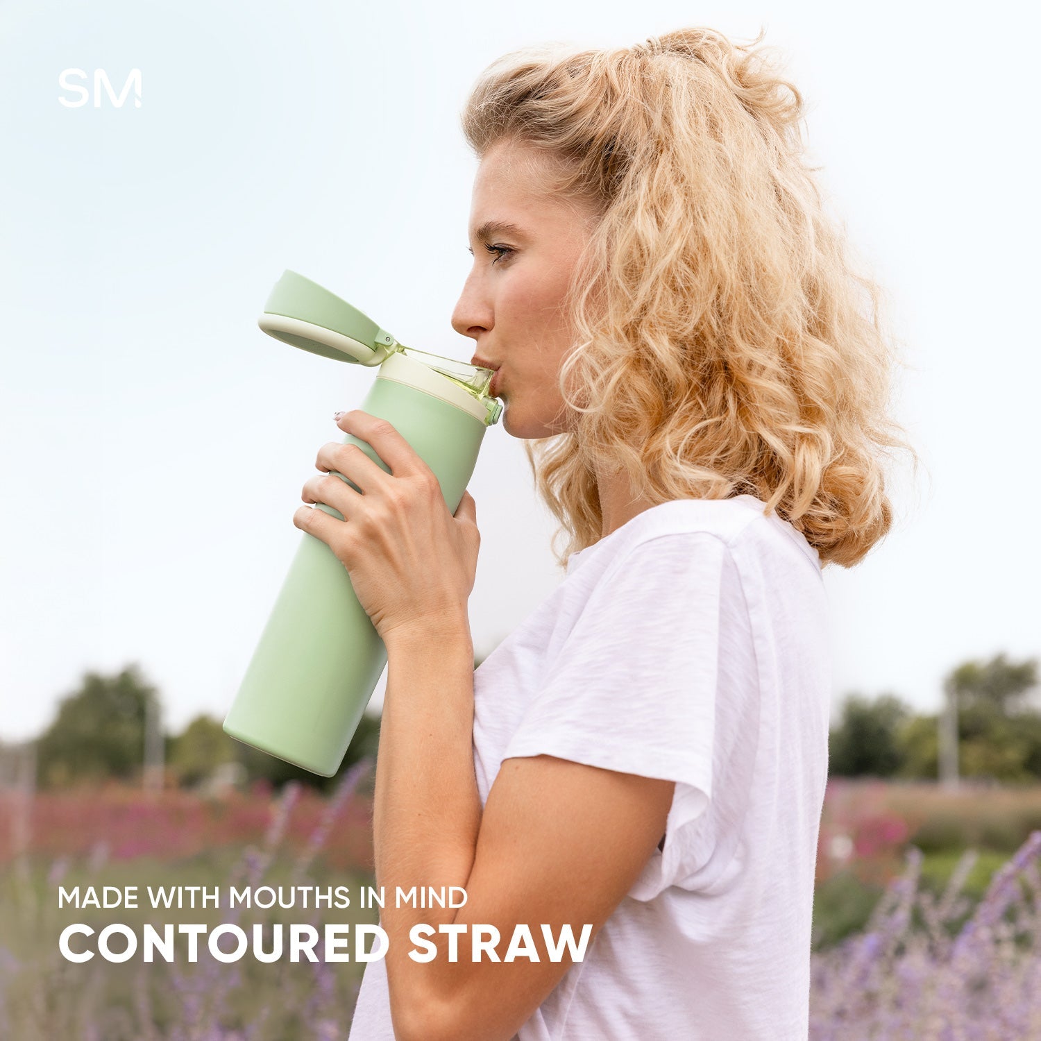 Woman with blonde hair drinking from a sage green contoured water bottle in a lavender field. White text reads "Made with mouths in mind" and "Contoured straw."