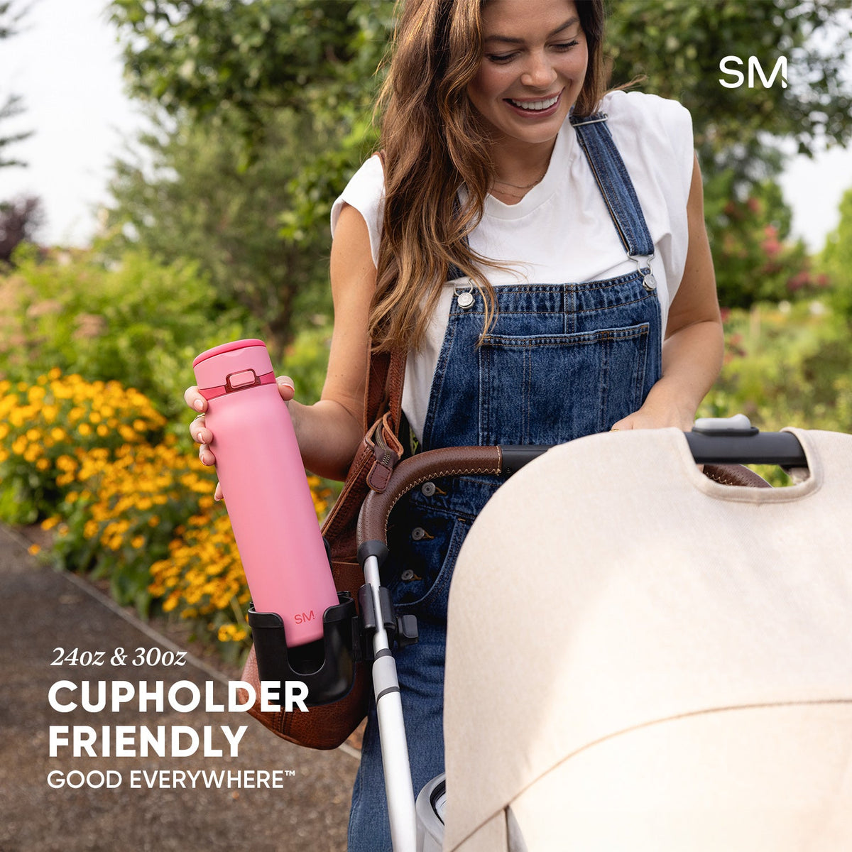Woman holding pink Simple Modern water bottle while pushing stroller in sunny garden. Bottle features cupholder-friendly design in 24oz and 30oz sizes. Text reads "Good Everywhere."