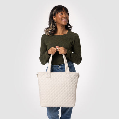 Cream quilted tote bag held by model wearing olive-green long-sleeved top and medium-wash blue jeans against neutral background