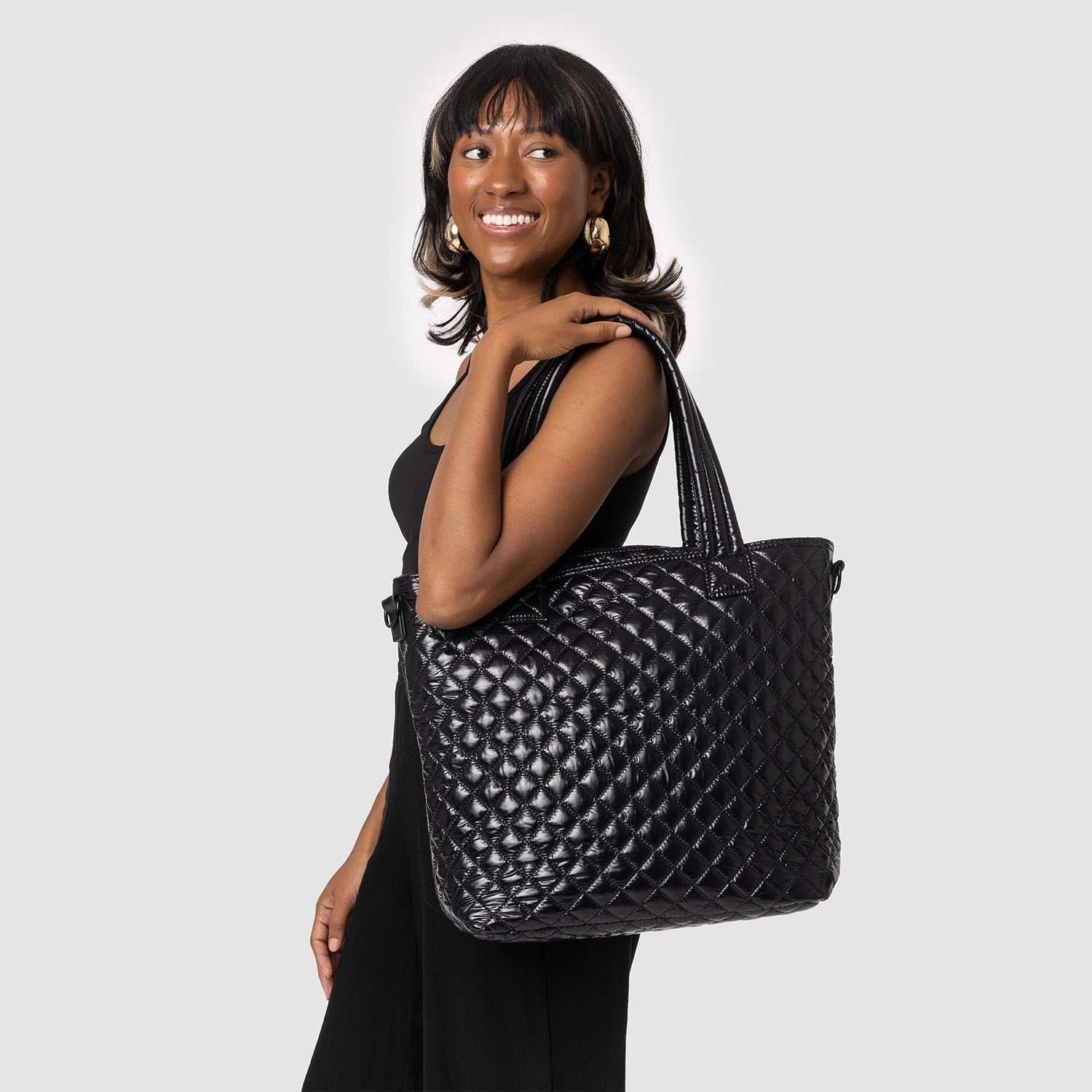 Woman wearing black outfit holding a black quilted tote bag with gold-toned hardware against a light gray background