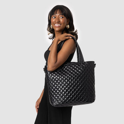 Woman wearing black outfit holding a black quilted tote bag with gold-toned hardware against a light gray background
