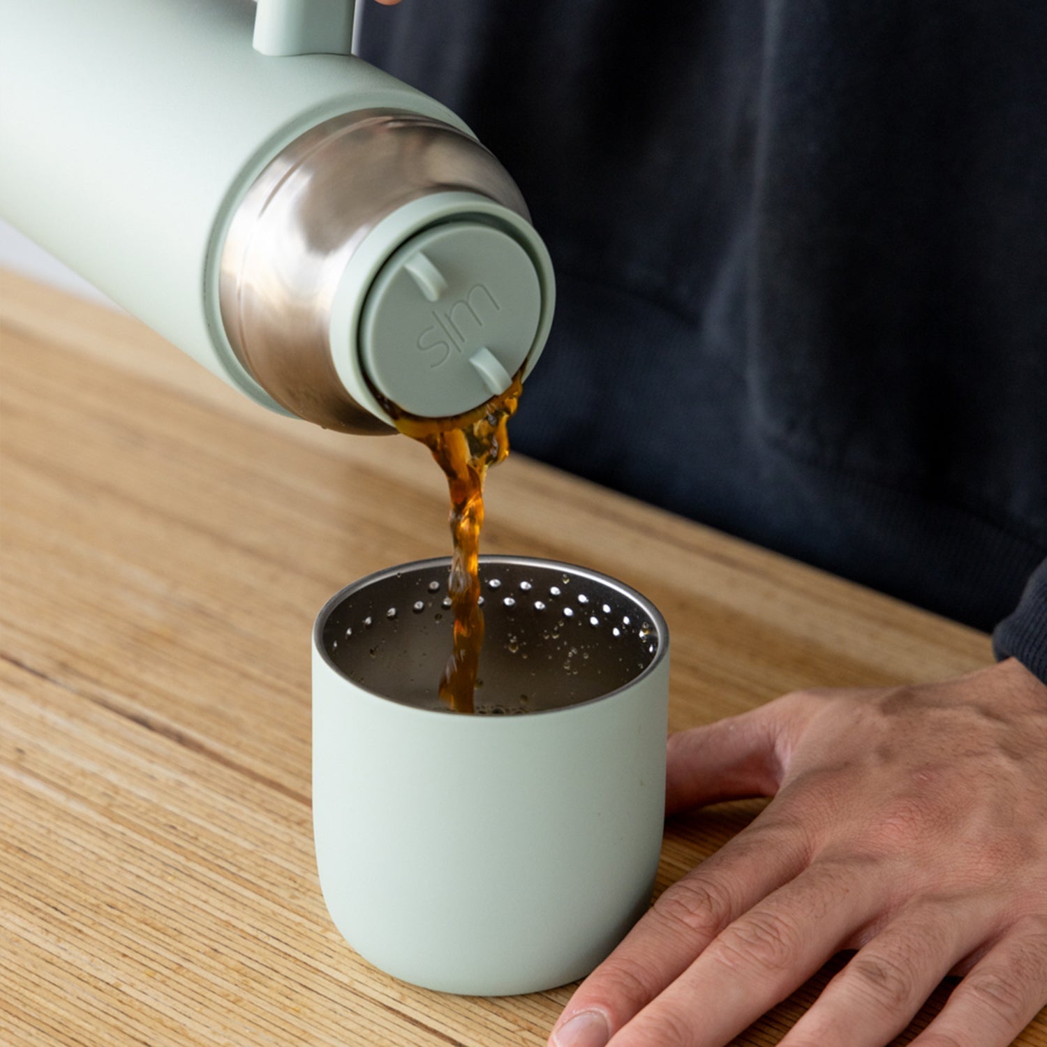 Mint green Simple Modern tumbler with lid pouring amber-colored coffee or tea into a matching cup with perforated strainer insert, held in a person's hand.