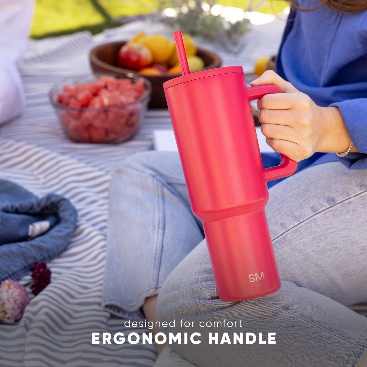 Coral-pink Trek Tumbler with ergonomic handle being held at an outdoor picnic, surrounded by picnic blanket, fruit bowl, and berries, with text "designed for comfort ERGONOMIC HANDLE"