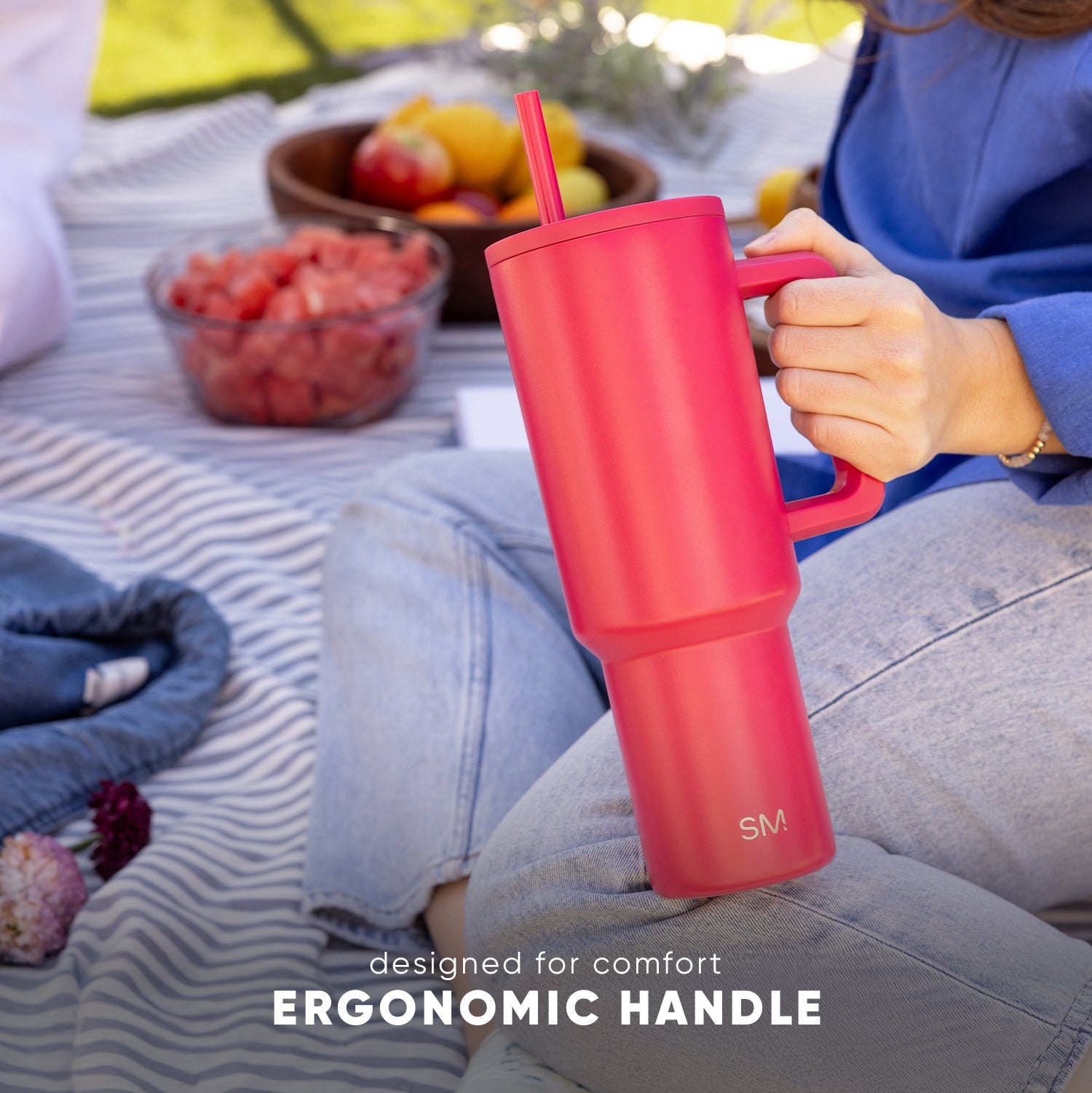 Coral-pink Trek Tumbler with ergonomic handle being held at an outdoor picnic, surrounded by picnic blanket, fruit bowl, and berries, with text "designed for comfort ERGONOMIC HANDLE"