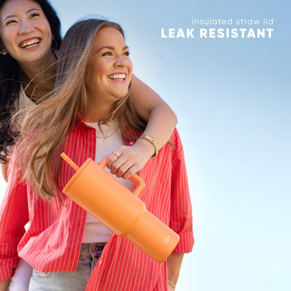Two smiling women outdoors holding a bright orange Trek Tumbler with matching straw lid against a blue sky background. Text reads "insulated straw lid" and "LEAK RESISTANT."