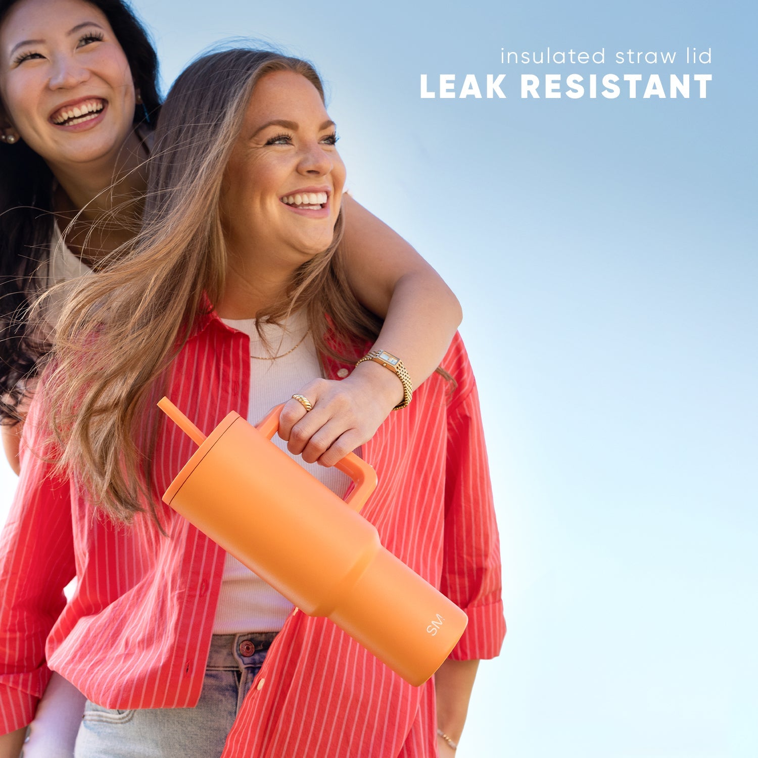 Two smiling women outdoors holding a bright orange Trek Tumbler with matching straw lid against a blue sky background. Text reads "insulated straw lid" and "LEAK RESISTANT."