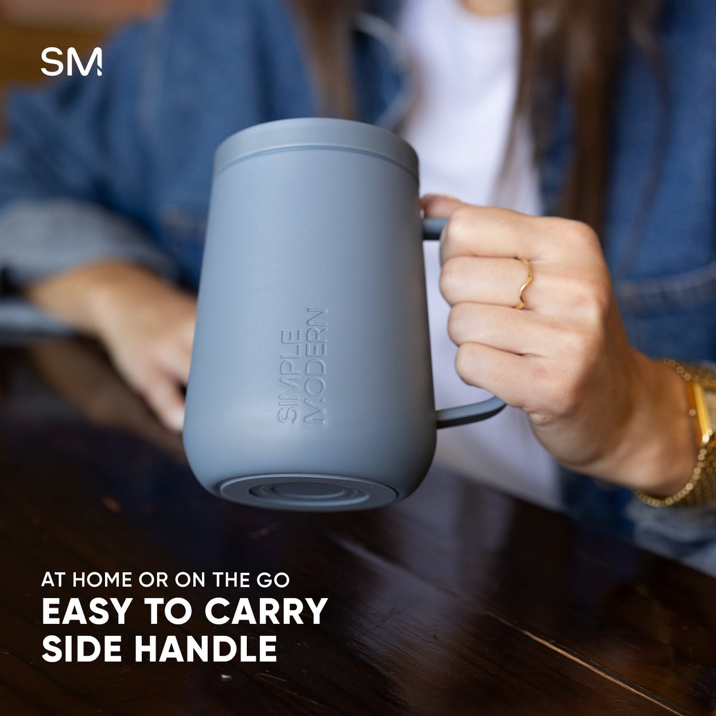Pale blue-gray Voyager360 mug with handle held by a hand, displayed against warm wood tones and navy clothing, showing product features and ergonomic design.