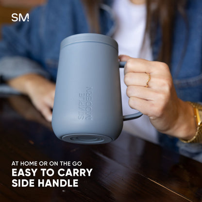 Pale blue-gray Voyager360 mug with handle held by a hand, displayed against warm wood tones and navy clothing, showing product features and ergonomic design.