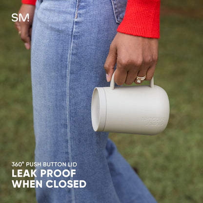 Cream-colored cylindrical mug held by person in light blue denim, demonstrating leakproof feature of Voyager360 drinkware.