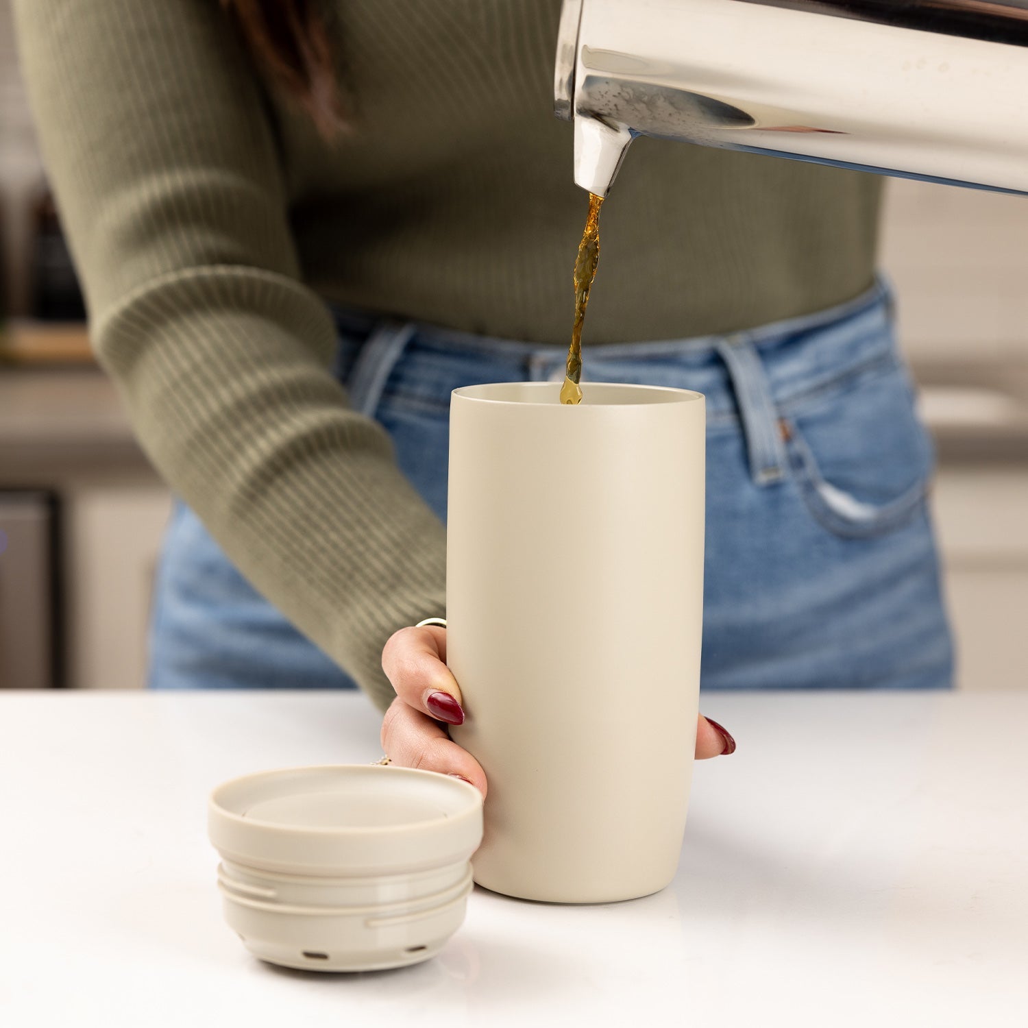 Cream-colored insulated drinkware displayed next to a person wearing a sage green ribbed sweater and blue denim, demonstrating leakproof design features.
