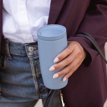 Pale blue tumbler held by person wearing deep burgundy jacket against soft blue and periwinkle background
