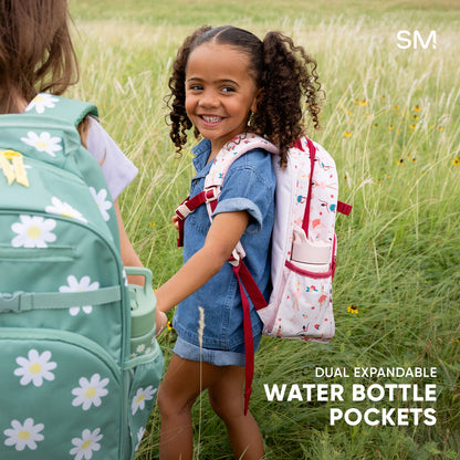 Fletcher Kids' Backpack in medium size with rainbow horizontal stripes, worn by a child on playground equipment. Features dual water bottle pockets, multiple zippered compartments, mesh panels, and orange handles.