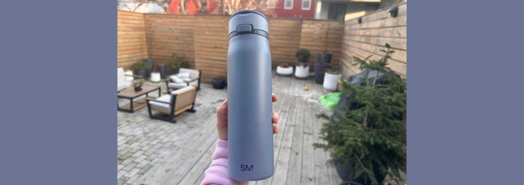 Person holding light blue Simple Modern insulated water bottle with flip-top lid on residential patio with wooden deck, fence, and potted plants in background.