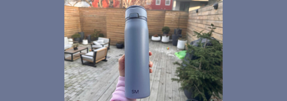 Person holding light blue Simple Modern insulated water bottle with flip-top lid on residential patio with wooden deck, fence, and potted plants in background.