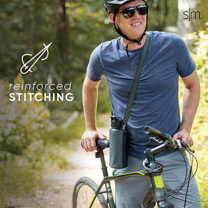 Man in blue-gray t-shirt and khaki pants standing with a sling-style bag across his body, holding a bicycle with black frame against green foliage background.